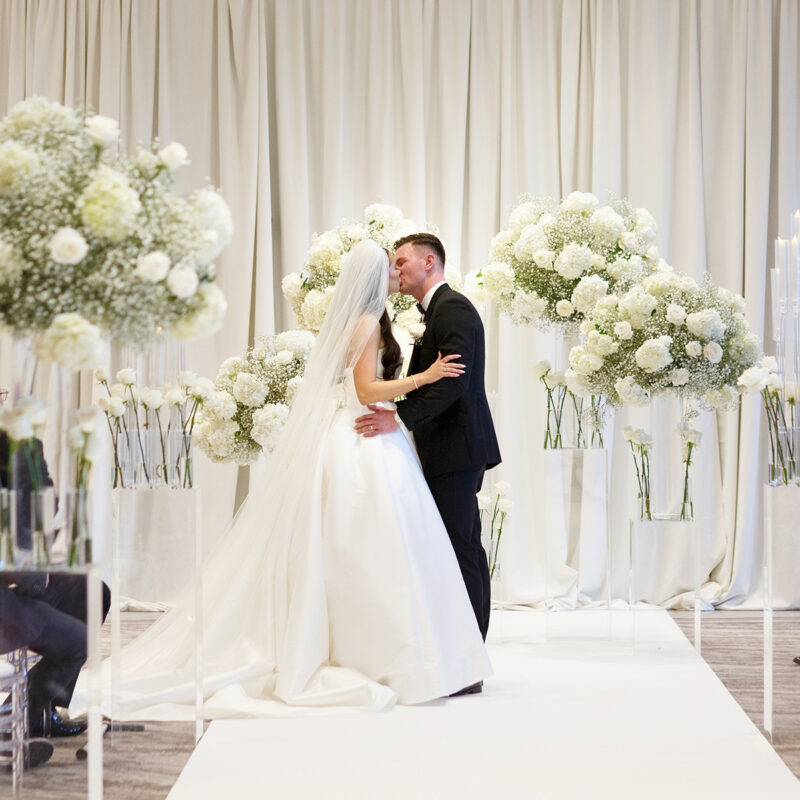 Bride and groom kissing on white aisle with floral plinths at Cameron House by Muse Styling